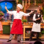 Theaterszene mit Schauspielern auf bunter Bühne beim Stück ‚La cuisine de ma cousine‘ – KI-Symbolbild